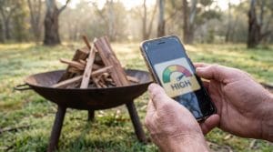 A weathered pair of hands holding a smartphone displaying a fire danger rating, framed against an unlit campfire of neatly stacked kindling and hardwood in a simple steel fire bowl on legs, early morning light filtering through eucalyptus trees in an Australian bush campsite, dew still on the grass, shallow depth of field with the phone sharp and the cold fire pit softly blurred behind, quiet and cautious mood, no text legible.