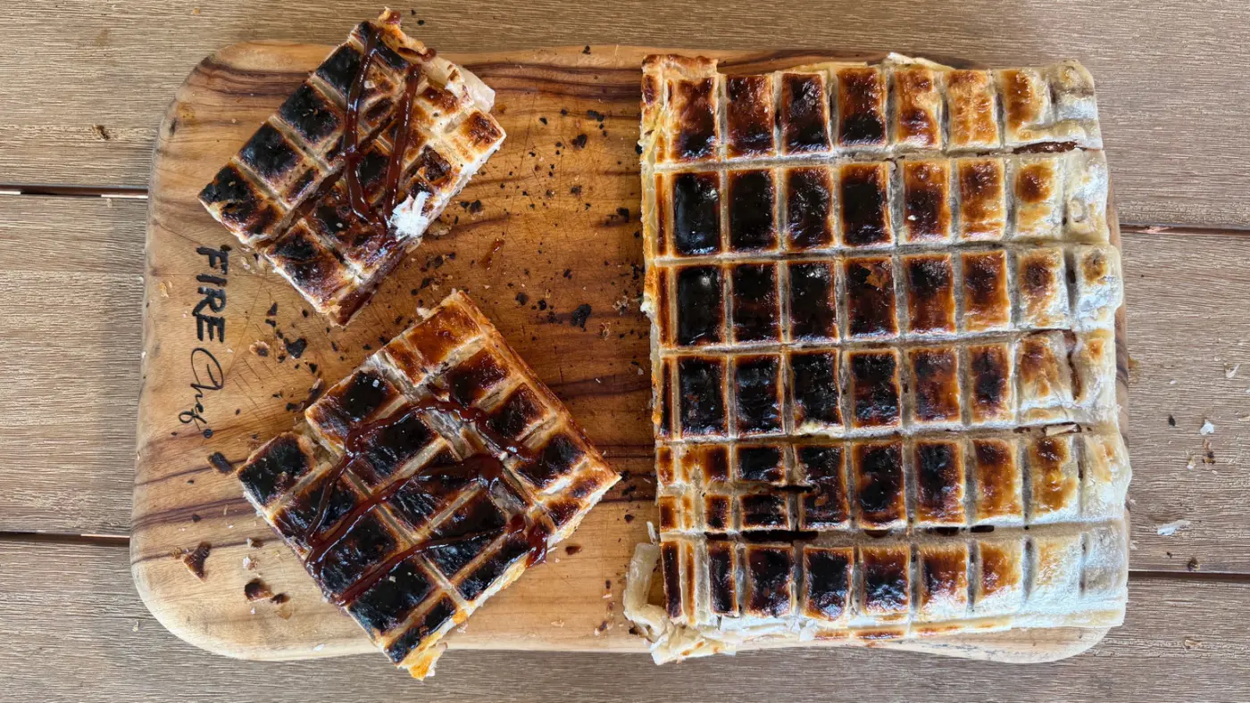 Rectangular puff pastry braai pie with dark grid marks, with two sauce-topped portions on a wooden board