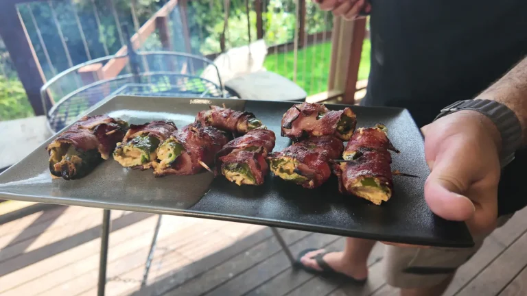 Bacon-wrapped green jalapeño portions with filling and toothpicks on a black and grey rectangular plate